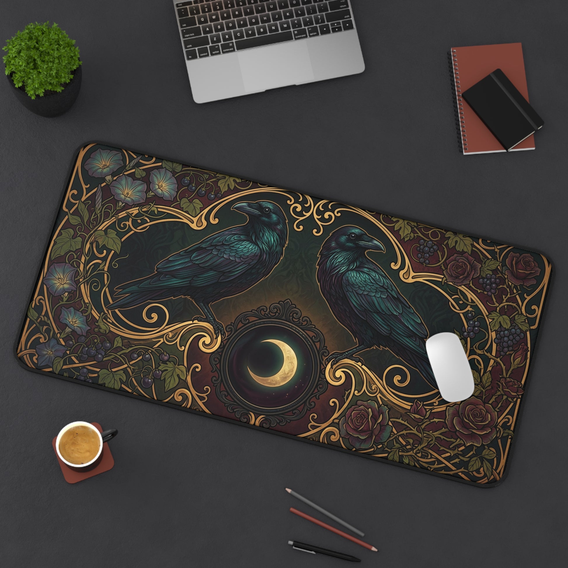 A large rectangular desk mat with an ornate raven and crescent moon design, shown on a dark gray desk with a laptop, notebook, plant, coffee cup, and pens around it.