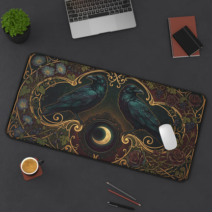 A large rectangular desk mat with an ornate raven and crescent moon design, shown on a dark gray desk with a laptop, notebook, plant, coffee cup, and pens around it.