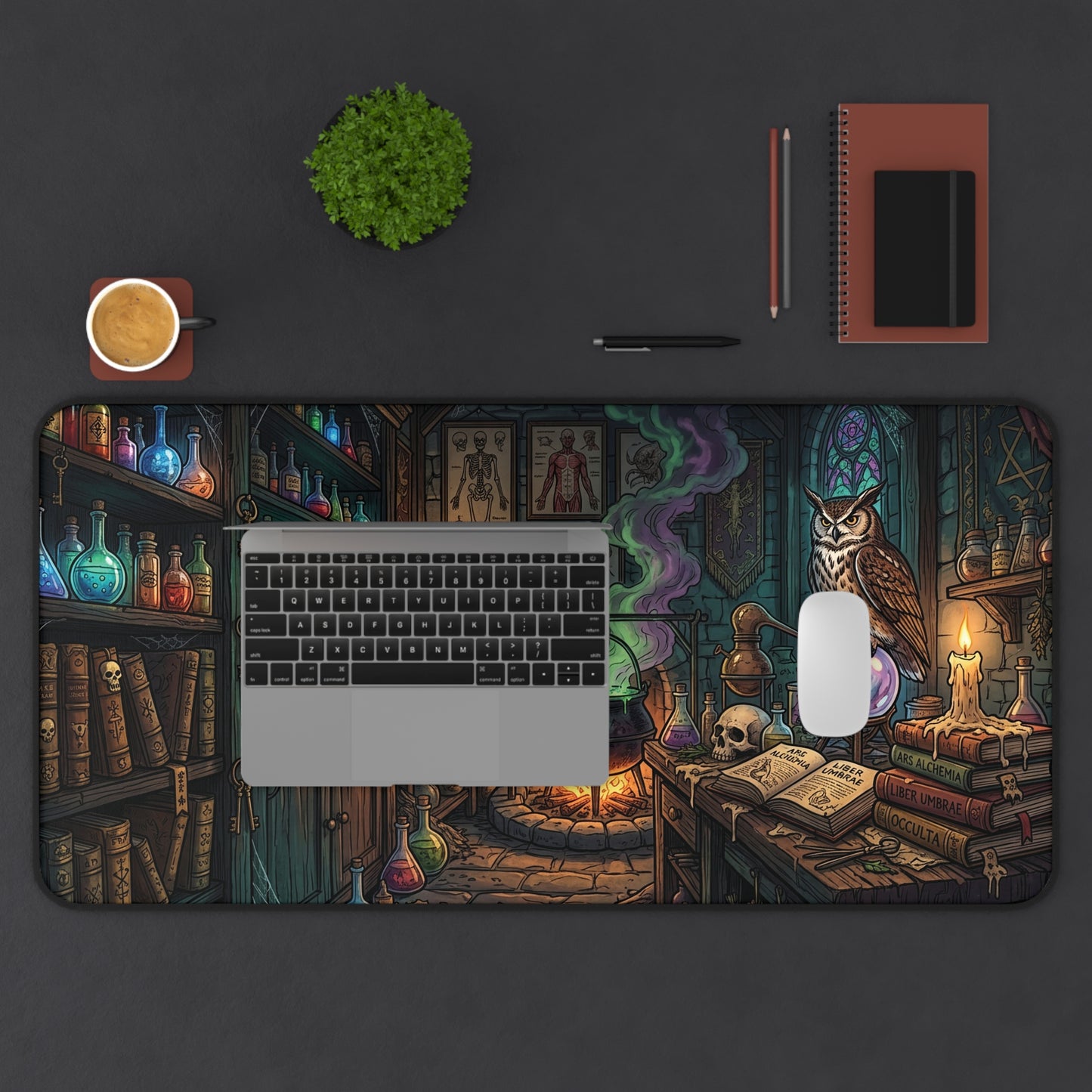 A large desk mat featuring a detailed wizarding library and alchemy lab illustration, styled on a dark gray desk with a laptop, mouse, notebook, plant, and coffee cup.