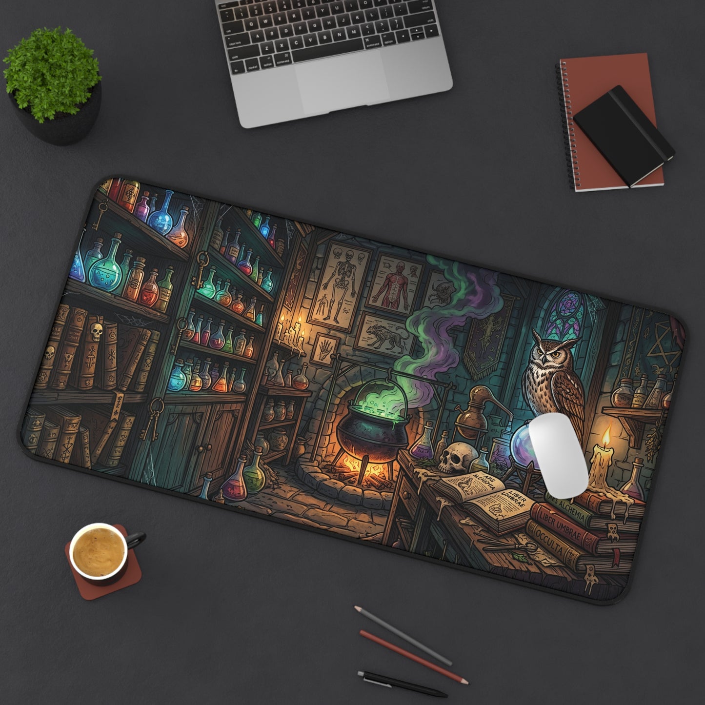 A large rectangular desk mat with a detailed fantasy illustration of a witch's library, shown on a dark gray desk with a laptop, coffee cup, and notebooks around it. The mat features an owl, cauldron, books, and glowing potions.