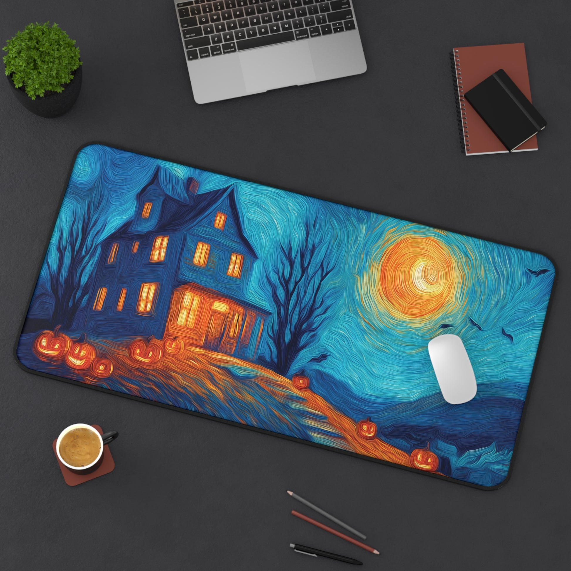 A large rectangular desk mat with a stylized Halloween night scene printed on it, shown on a dark tabletop with a laptop, notebook, plant, coffee cup, and pens around it. The mat features a blue swirling sky, a glowing orange moon, jack-o'-lanterns, a haunted house with warm windows, and bare trees.