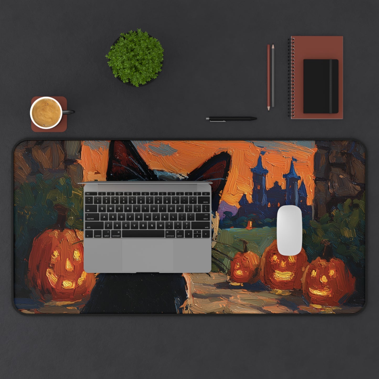A large rectangular desk mat with a Halloween-themed painting of jack-o'-lanterns, a witch's hat, and a haunted castle, shown on a dark gray desk with a laptop, mouse, notebook, and plant.