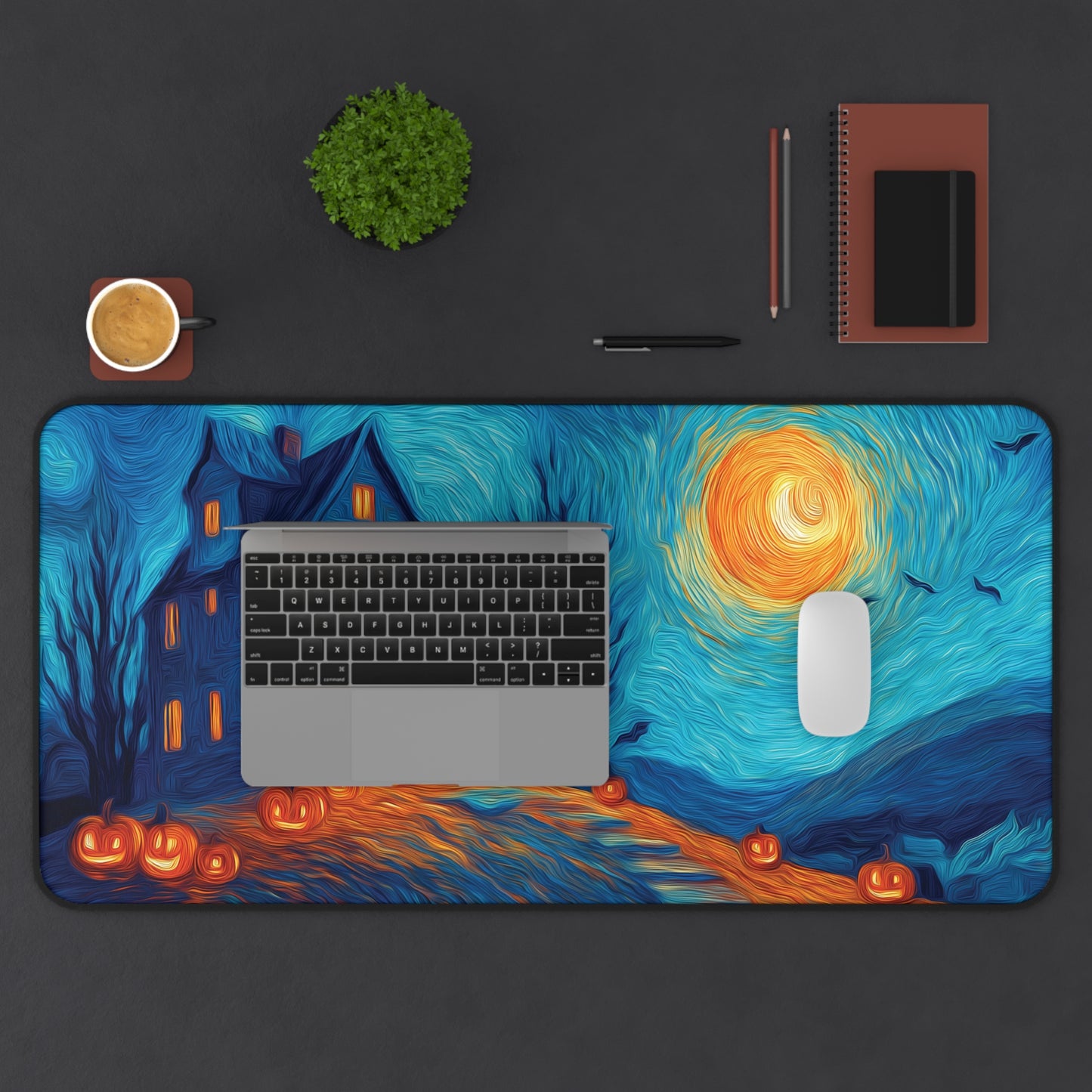 A large desk mat with a stylized Van Gogh-inspired Halloween scene, featuring a haunted house, glowing pumpkins, swirling blue sky, and an orange moon, shown on a dark desk with a laptop, mouse, notebook, and other desk accessories.