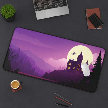 A large rectangular desk mat with a purple and black Halloween landscape design featuring a haunted house, full moon, and witch on a broomstick, shown on a dark gray desk with a laptop, plant, notebook, coffee cup, and pens around it.
