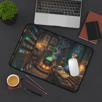 A large desk mat with a detailed dark fantasy illustration of a witch's laboratory, styled in a flat-lay workspace scene with a laptop, coffee cup, pencils, and notebooks on a dark gray surface.