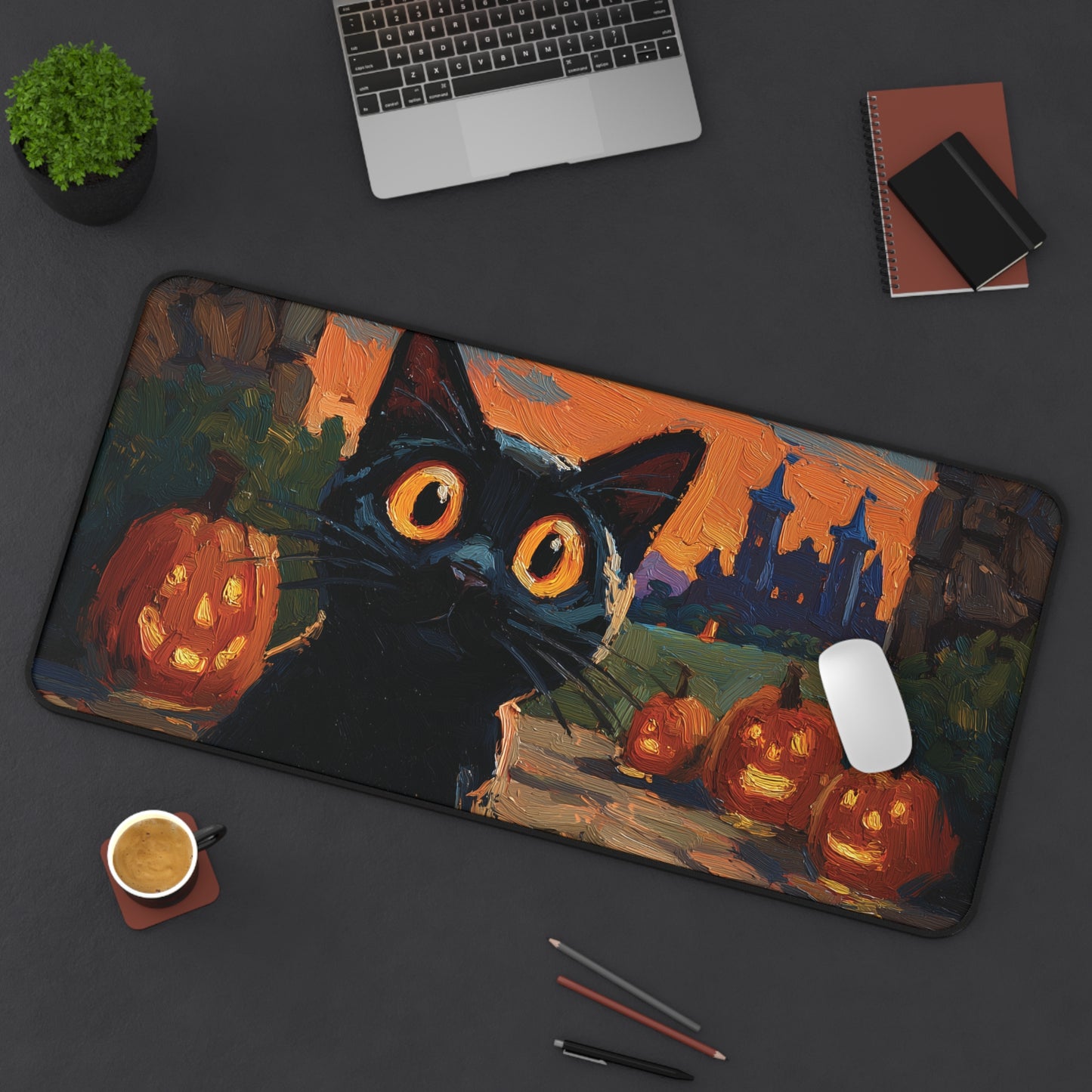 A large rectangular desk mat with a Halloween-themed painting of a black cat, glowing jack-o'-lanterns, and a castle at sunset, shown on a dark gray desk with a laptop, notebook, plant, coffee cup, and pens around it.