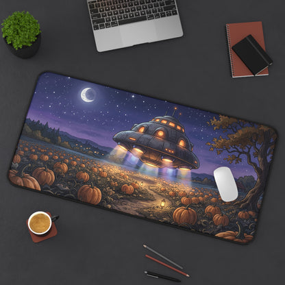 A large rectangular desk mat with a whimsical Halloween night scene, displayed on a dark gray desk with a laptop, coffee cup, notebook, and plant. The desk mat features an illustration of a flying saucer hovering over a pumpkin field under a starry sky with a crescent moon.