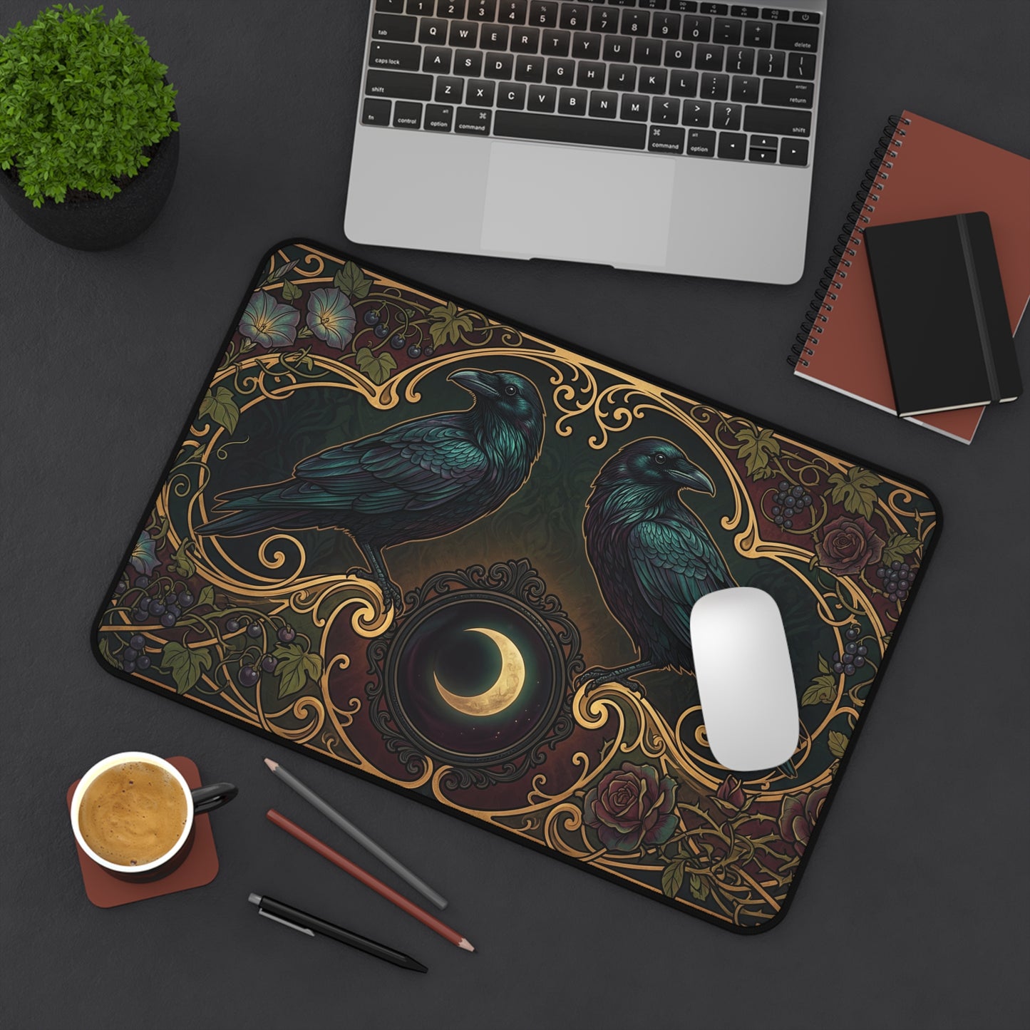 A rectangular desk mat with an ornate raven and crescent moon design, styled on a dark gray desk with a laptop, mouse, coffee cup, and notebooks.