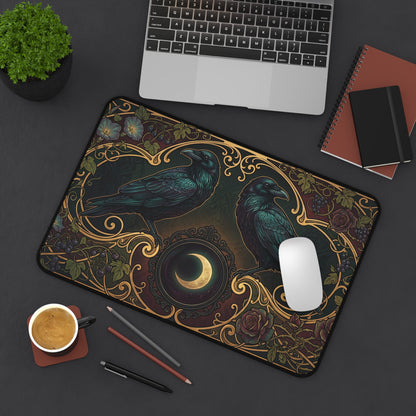 A rectangular desk mat with an ornate raven and crescent moon design, styled on a dark gray desk with a laptop, mouse, coffee cup, and notebooks.