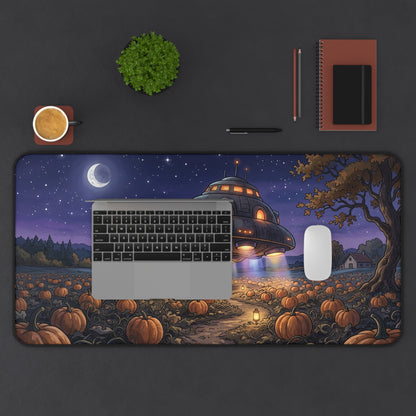 A large rectangular desk mat with a Halloween night scene print, shown on a dark gray desk with a laptop, mouse, notebook, plant, and coffee cup. The mat features a pumpkin field, a tree, a UFO, and a crescent moon.