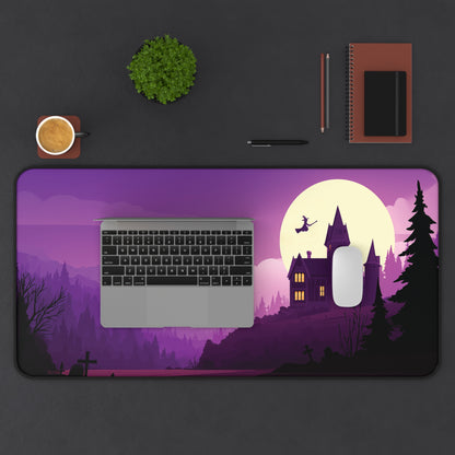 A large rectangular desk mat with a Halloween-themed illustration, shown on a dark gray desk with a laptop, mouse, notebook, plant, and coffee cup. The mat features a purple and black landscape with a castle, full moon, and witch silhouette.